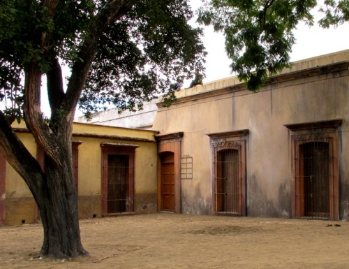 Dirt sidewalk and aged looking buildings and tree.