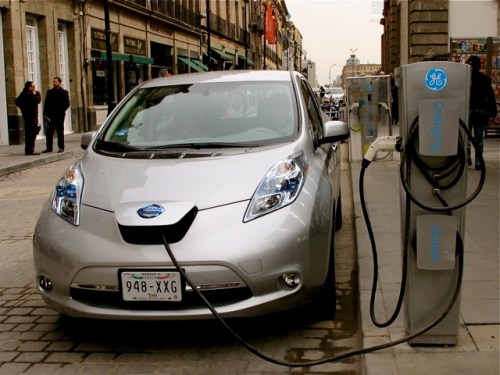 Silver Nissan electric car getting charged at charging station in the street.