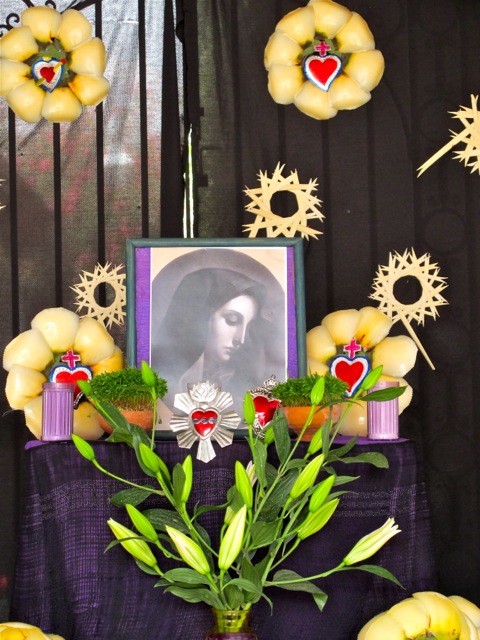 Photo on altar of La Virgen de Dolores with vase of lilies in forground and palm "flowers" in background