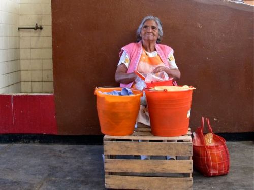 White-haired woman sitting behind two plastic buckets full of tamales
