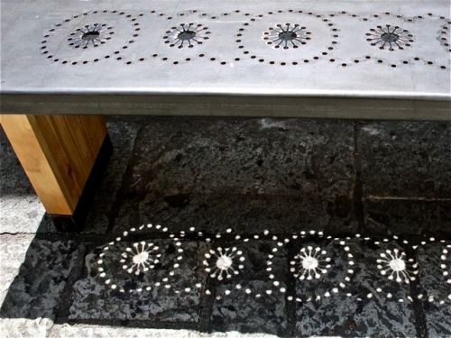 Bench with metal seat with punched design, which is mirrored in the shadow on the paving stones.