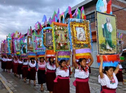 Young women wearing dark red wool wrap skirts and embroidered white blouses, carry large canastas on their heads